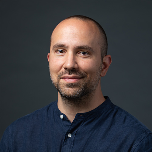 A headshot of CS assistant professor Evgenios Kornaropoulos, wearing a dark blue button-up shirt, smiling slightly against a dark gray background.